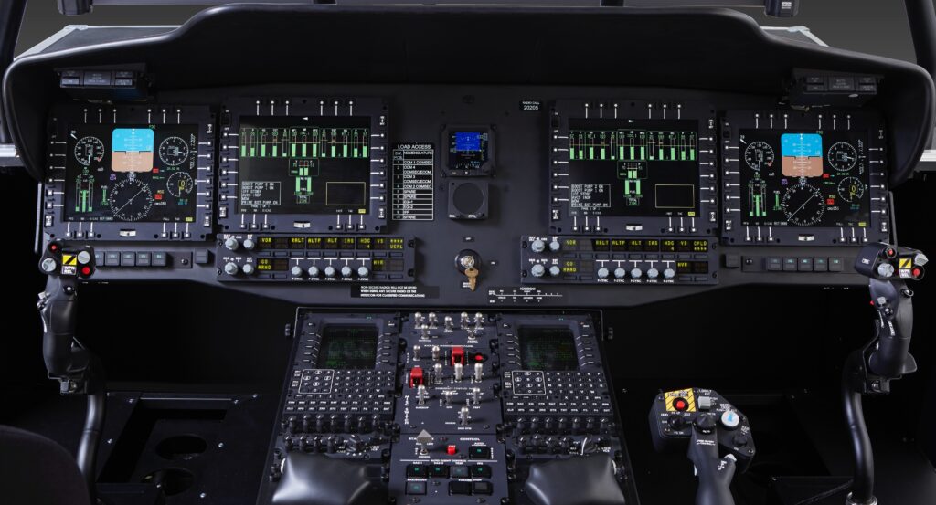 Cockpit view showing an array of digital flight displays, control panels with numerous buttons and switches, dual joysticks, and illuminated instrument panels for piloting an aircraft.