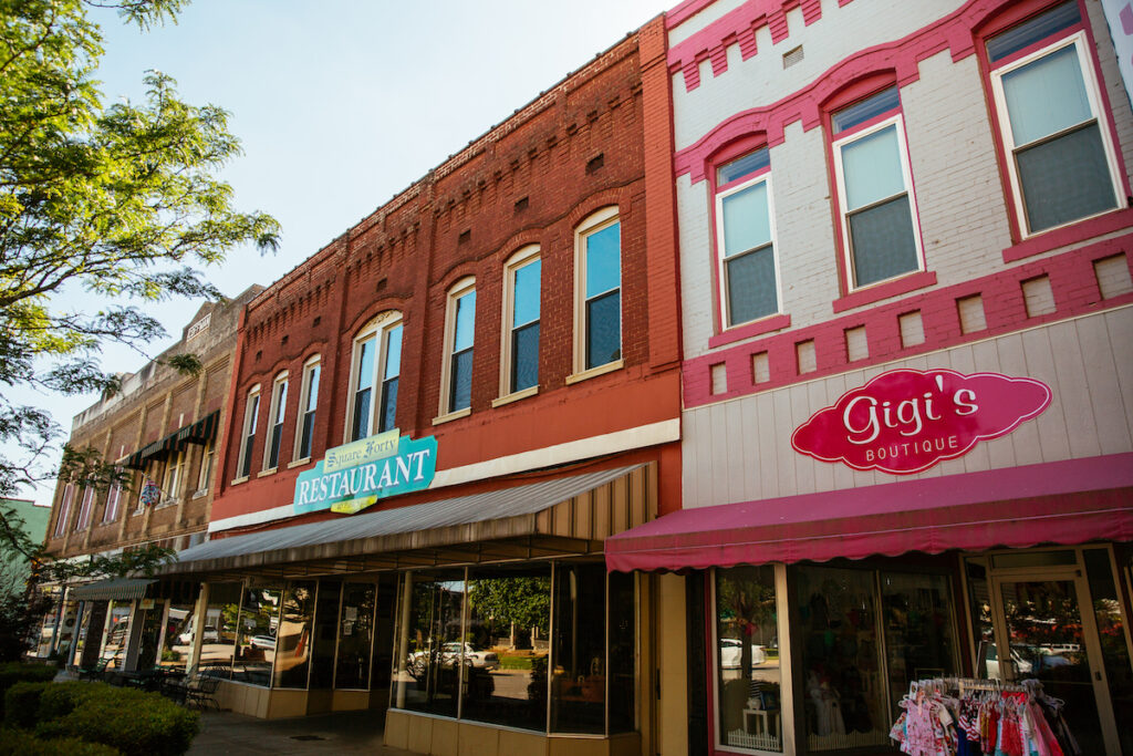 A row of colorful downtown storefronts, including a restaurant with a turquoise sign and Gigi’s Boutique with a pink sign, on a sunny day. Clothes are displayed on racks outside the boutique.