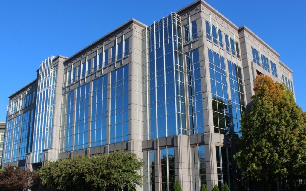 A modern office building with large, reflective glass windows and concrete trim, surrounded by green trees, under a clear blue sky.