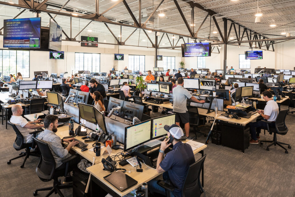 A busy, modern office with many people working at desks with multiple monitors. Some employees are seated, others are standing, and large screens hang from the ceiling displaying information. The space is bright with natural light.