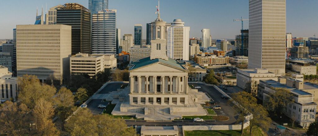 Aerial view of a neoclassical government building surrounded by modern skyscrapers and city infrastructure under clear skies. Trees and parked cars are visible in the foreground.