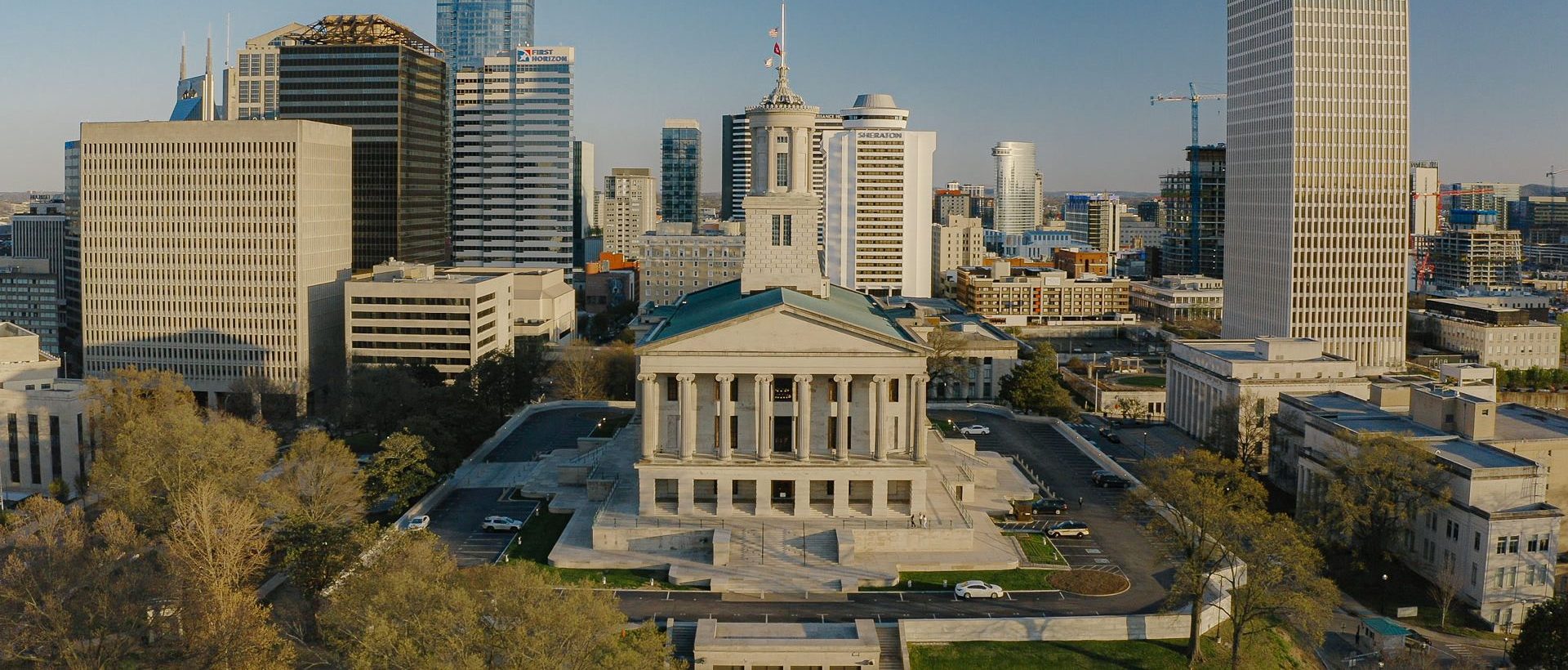 Aerial view of a neoclassical government building surrounded by modern skyscrapers and city infrastructure under clear skies. Trees and parked cars are visible in the foreground.