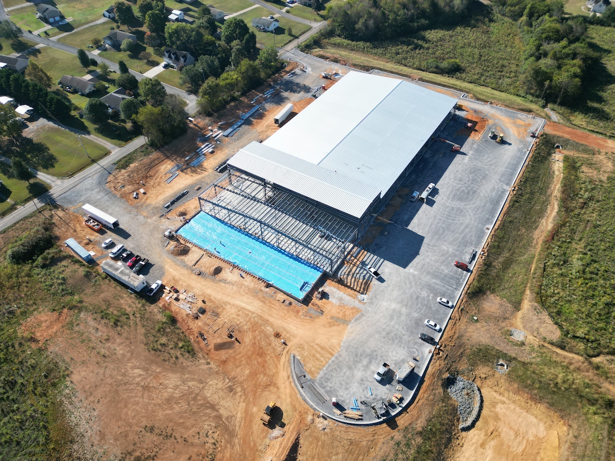 Aerial view of a large building under construction on a cleared lot, with steel framing and blue material visible. Surrounding the site are vehicles, equipment, and nearby green residential areas and trees.