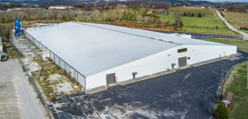 Aerial view of a large, white industrial warehouse with a metal roof, surrounded by paved parking areas and greenery, located in a rural setting with hills and scattered buildings in the background.