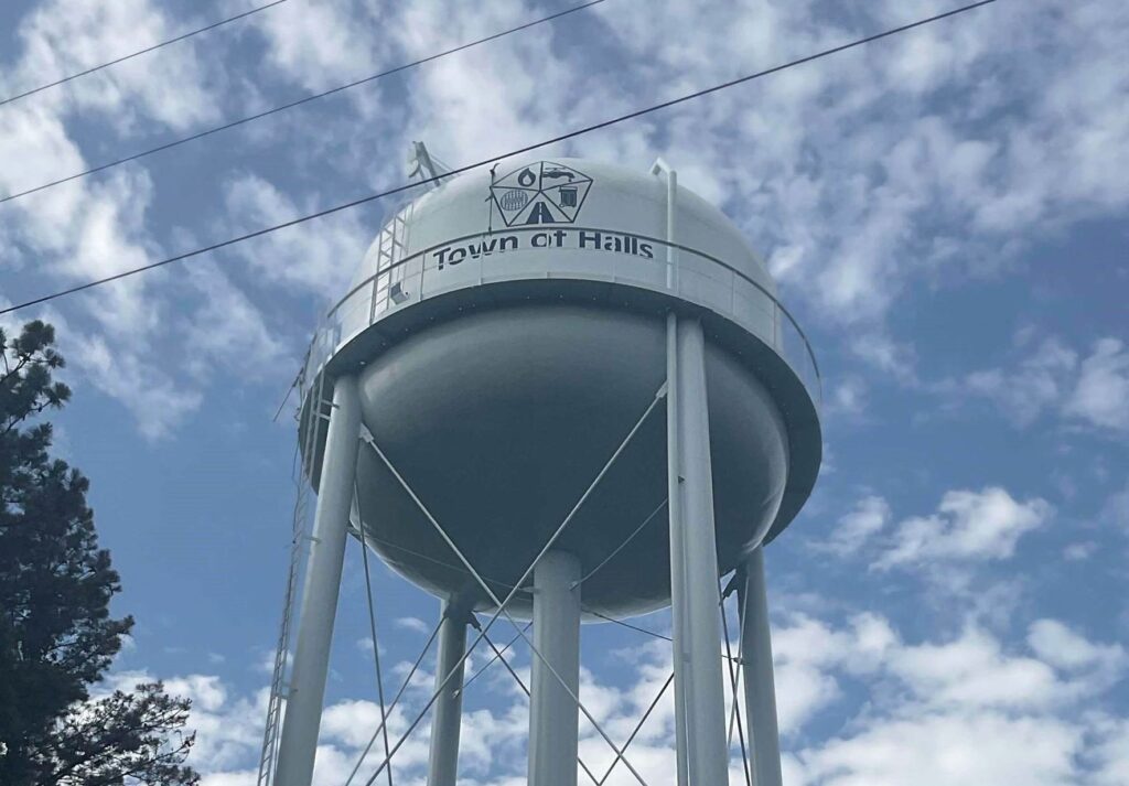 A tall white water tower with "Town of Halls" and a logo printed on the tank, stands against a partly cloudy blue sky, with tree branches visible on the left.