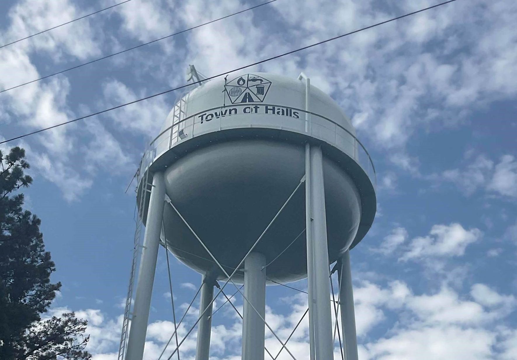A tall white water tower with "Town of Halls" and a logo printed on the tank, stands against a partly cloudy blue sky, with tree branches visible on the left.
