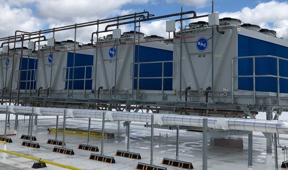 Four large industrial cooling towers with blue panels and BAC logos are installed on a rooftop. Pipes and metal railings run along the structure under a partly cloudy sky.
