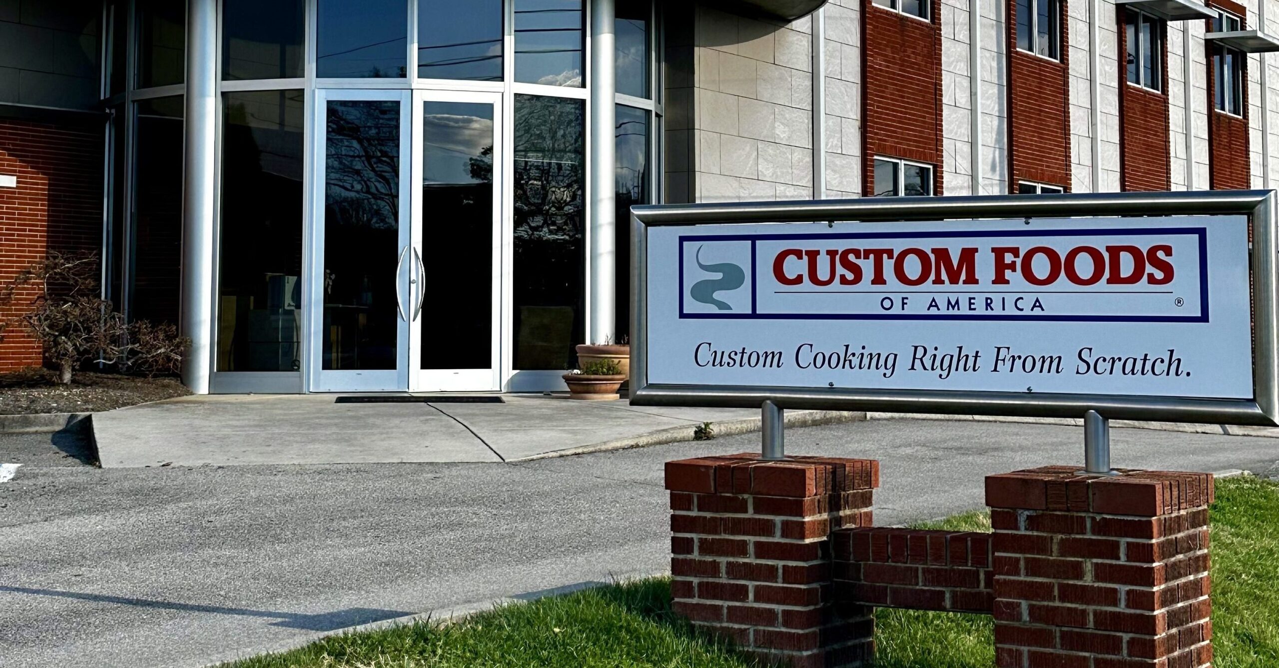 A sign reading "Custom Foods of America: Custom Cooking Right From Scratch" stands outside a modern building with glass doors and brick walls.