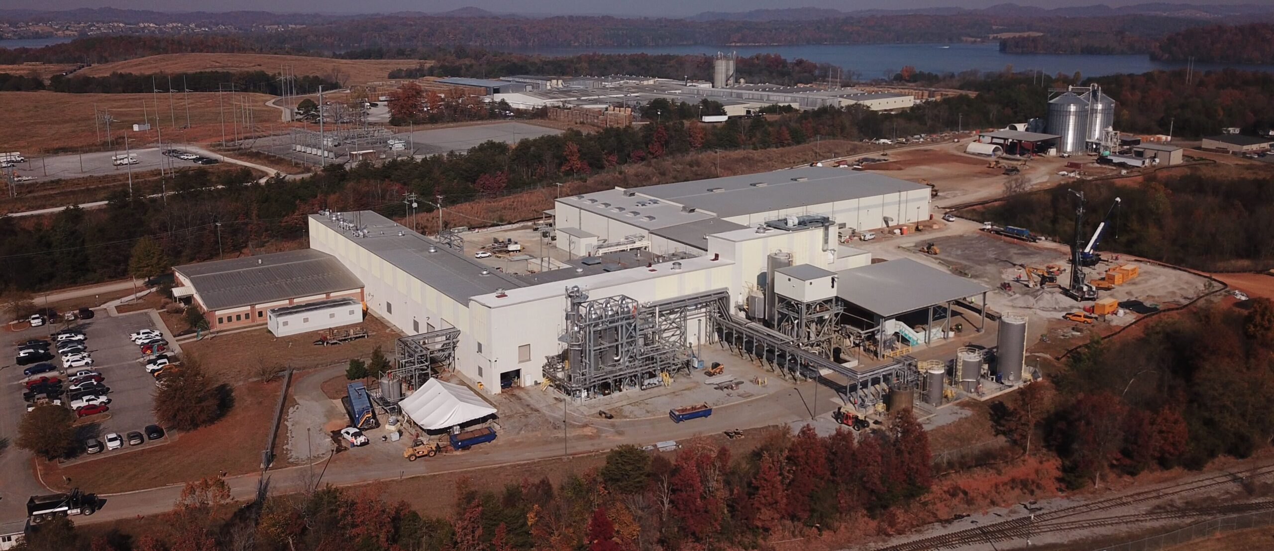 Aerial view of a large industrial facility surrounded by autumn trees, parking lots with cars, storage silos, and nearby water, with hills and additional buildings in the background.