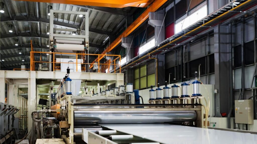 A large industrial machine processes flat sheets of material inside a spacious, well-lit factory with metal beams, overhead cranes, and control panels visible.
