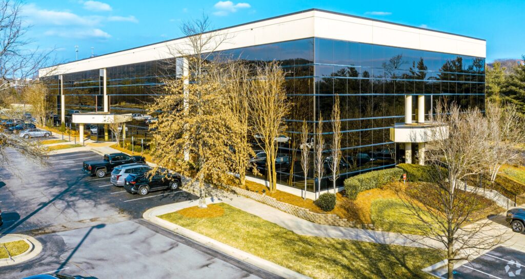 Modern office building with large reflective glass windows, surrounded by trees, parked cars, and a landscaped lawn under a blue sky.