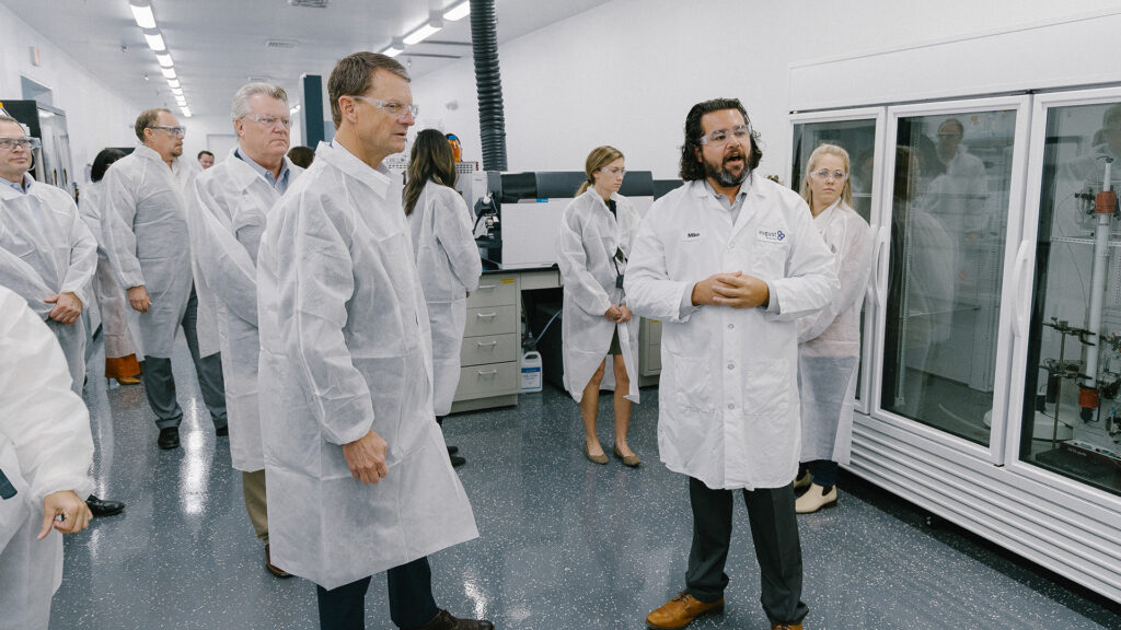A group of people wearing white lab coats and hairnets stand in a laboratory, listening to a man speaking in front of lab equipment and glass refrigerators. The setting appears clean and organized.