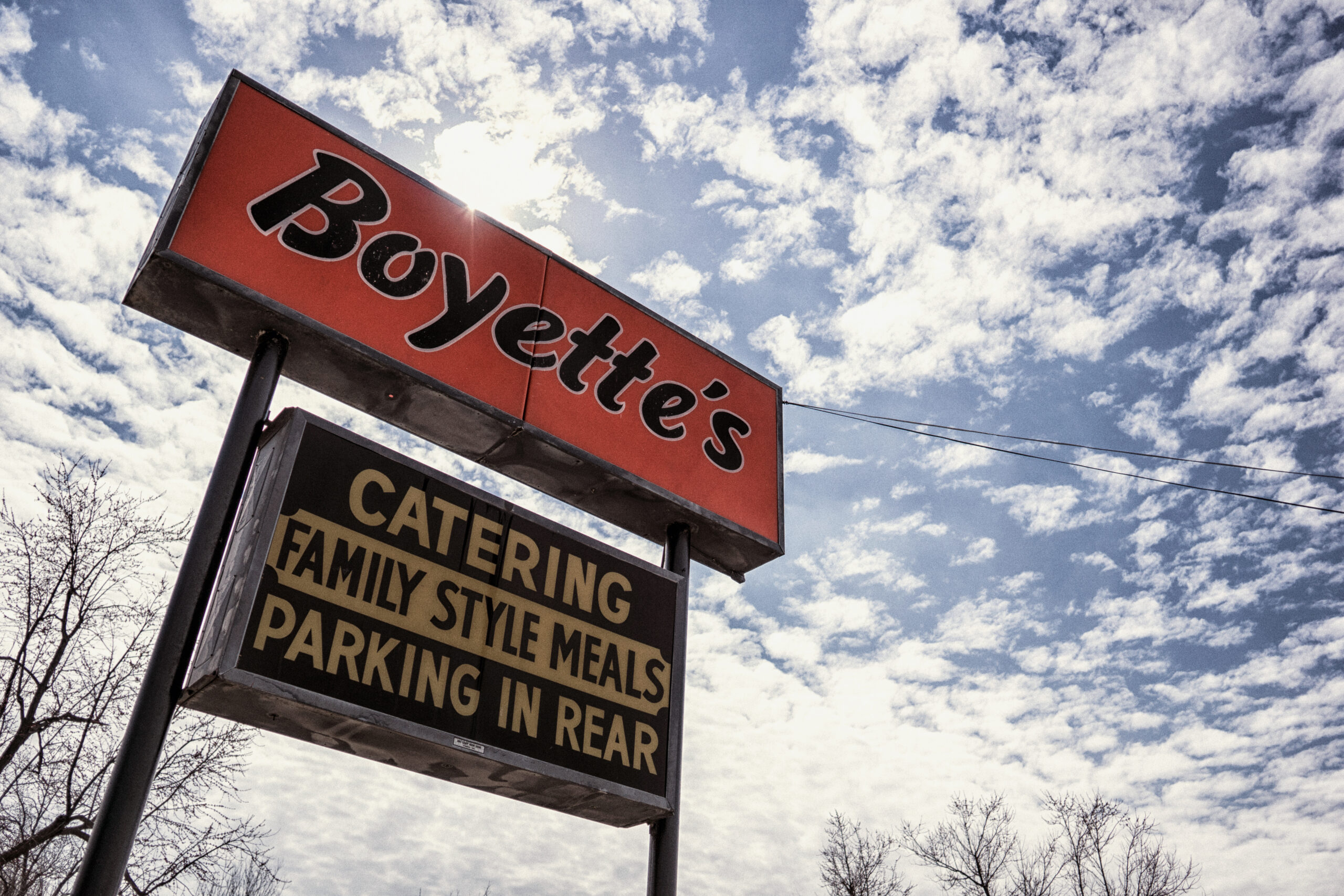 A red and black sign with "Boyette's" above a black and yellow sign reading "Catering Family Style Meals Parking in Rear" stands against a blue sky with scattered clouds and bare tree branches.