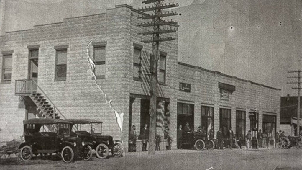 A black and white vintage photo of a two-story brick building, with early 20th-century cars parked outside. Several people stand on the sidewalk near utility poles, and an exterior stairway is visible on the left side.