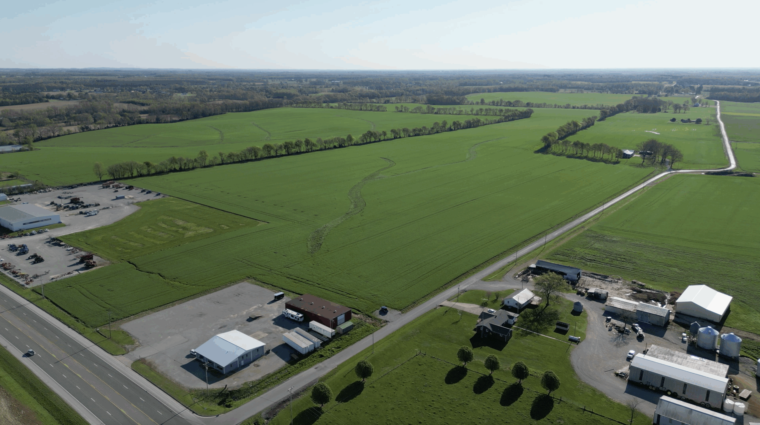 Aerial view of a rural landscape with green fields, scattered buildings, a few houses, roads, and a parking lot under a clear, blue sky. A line of trees borders the fields in the distance.