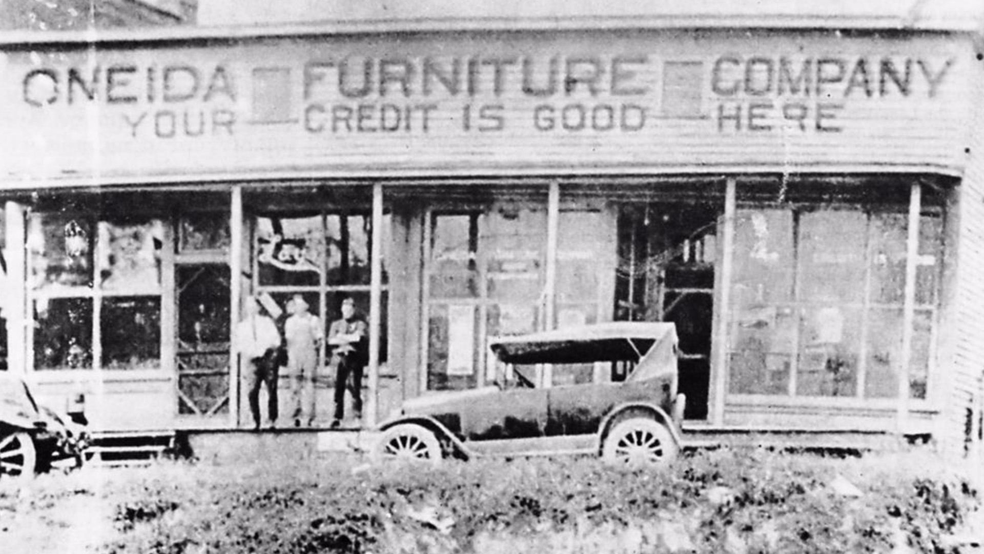 Black-and-white photo of the Oneida Furniture Company storefront with a sign reading “Your Credit is Good Here.” Four people stand in front of the store next to an old-fashioned car. Large windows display furniture inside.