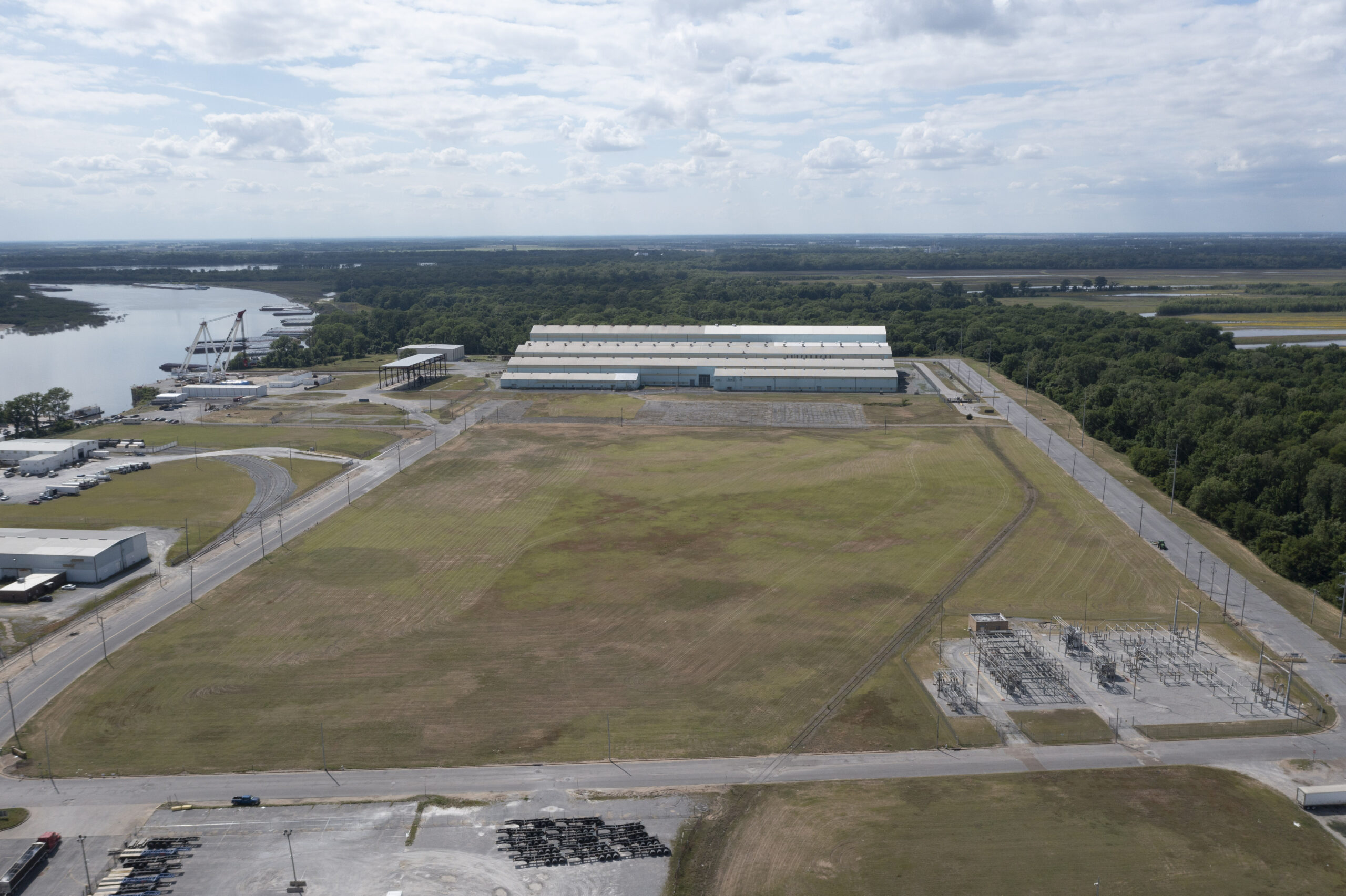 A large industrial complex with multiple warehouse buildings surrounded by a vast grassy field, near a river and wooded area under a partly cloudy sky. Power lines and utility structures are visible in the foreground.