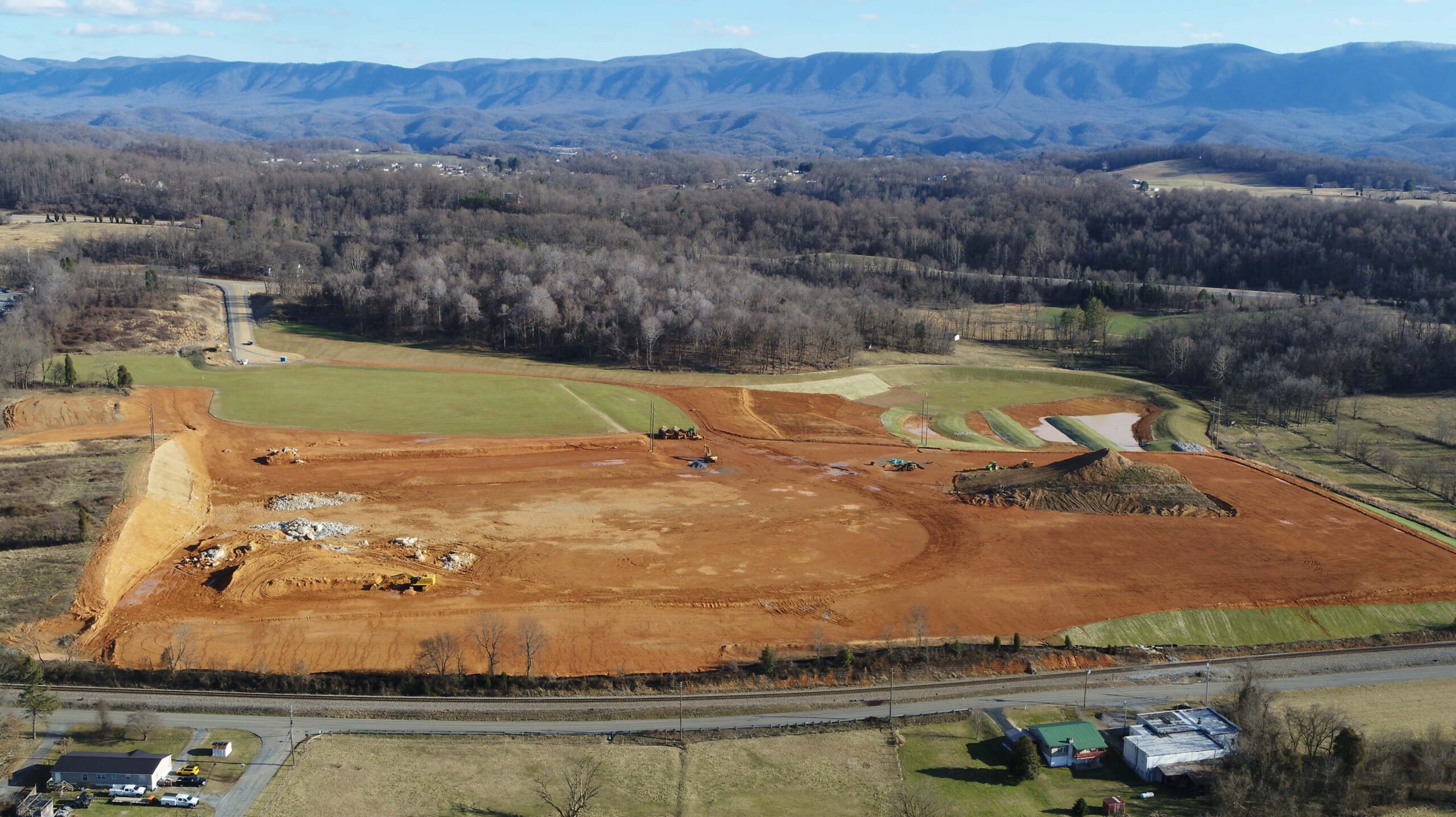 A large construction site with cleared red earth and machinery sits in a rural area, surrounded by fields, trees, and distant mountains under a blue sky.