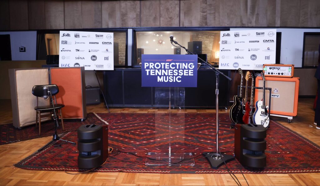 A clear podium with a “Protecting Tennessee Music” sign stands on a patterned rug, flanked by guitars, amplifiers, a microphone, speakers, and a display of sponsor logos in a recording studio.
