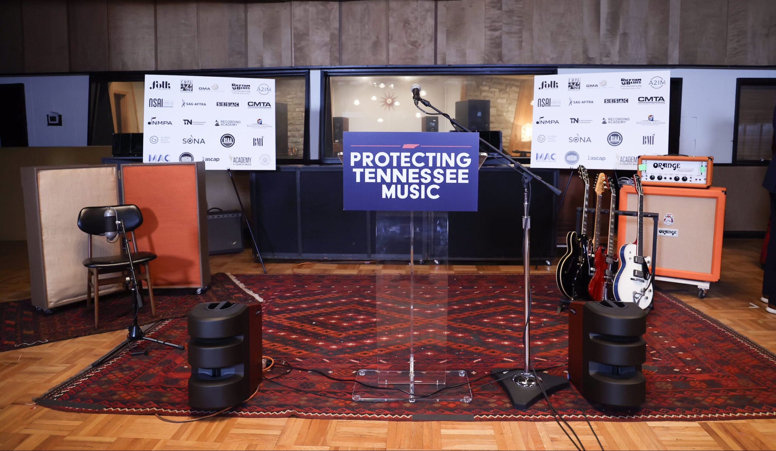 A clear podium with a “Protecting Tennessee Music” sign stands on a patterned rug, flanked by guitars, amplifiers, a microphone, speakers, and a display of sponsor logos in a recording studio.