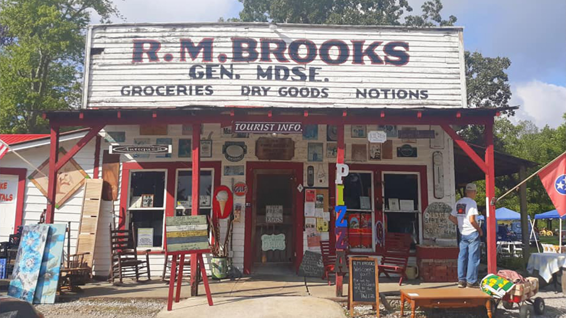A lively parade moves down a city street lined with tall buildings and crowds of people, many wearing green. Street signs and shops are visible, and the sky is partly cloudy.