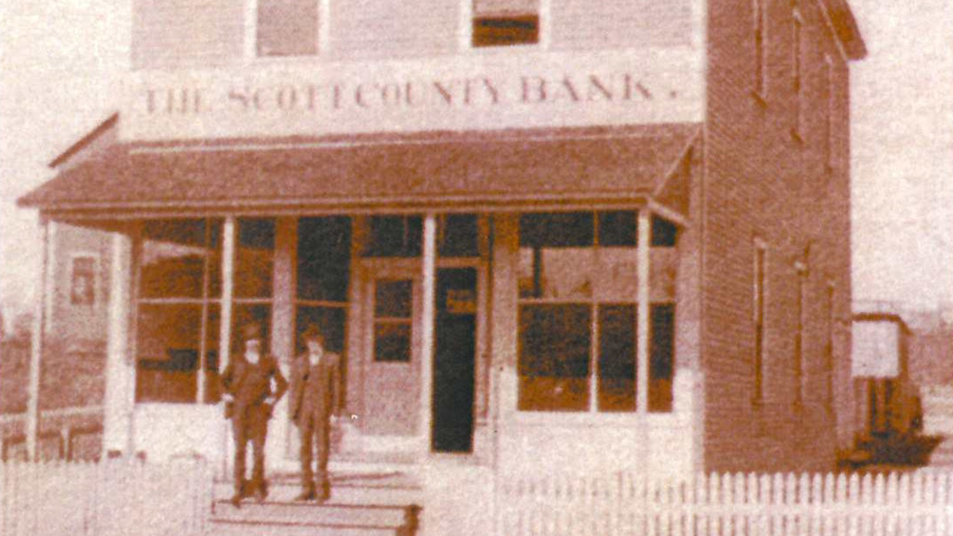 Sepia-toned photo of two men in suits standing in front of a two-story brick building with a porch and a sign reading “The Scott County Bank.” A white picket fence runs along the front.