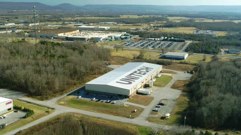 Aerial view of an industrial area with a large warehouse labeled "UNITECH" on the roof, surrounded by roads, trees, and several other buildings and parking lots in a rural landscape.