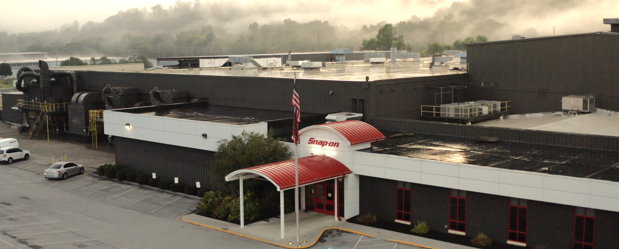 A large industrial building with a red-and-white "Snap-on" sign above the entrance, an American flag, parked cars, and mist rising from trees in the background. The roof and pavement appear wet from recent rain.