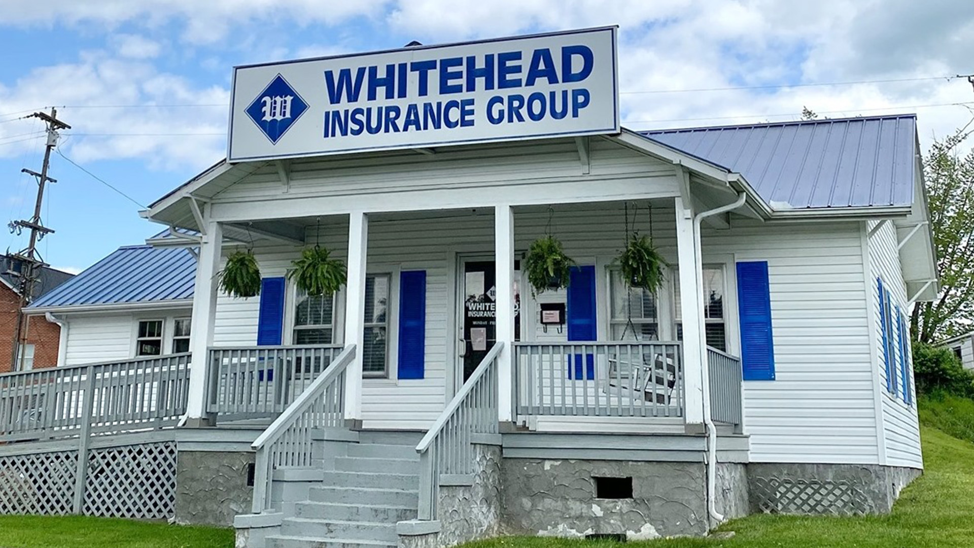 A small white building with blue shutters and a blue metal roof houses the Whitehead Insurance Group, as shown by a large sign above the entrance. The porch has hanging plants and steps leading up to the door.