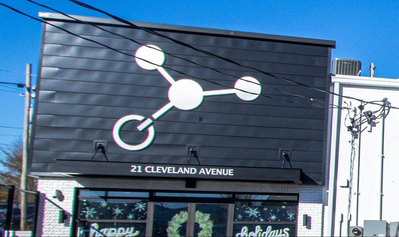 A black building facade features a large white molecular structure graphic and the address "21 Cleveland Avenue" above the entrance. There are holiday decorations visible in the windows and electrical wires overhead.
