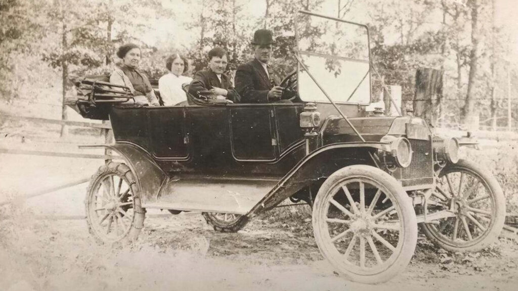 Four people sit in an old-fashioned, open-top car on a dirt road. Two women are in the back seat, while a man in a hat and another woman sit in front. Trees and a fence are visible in the background.