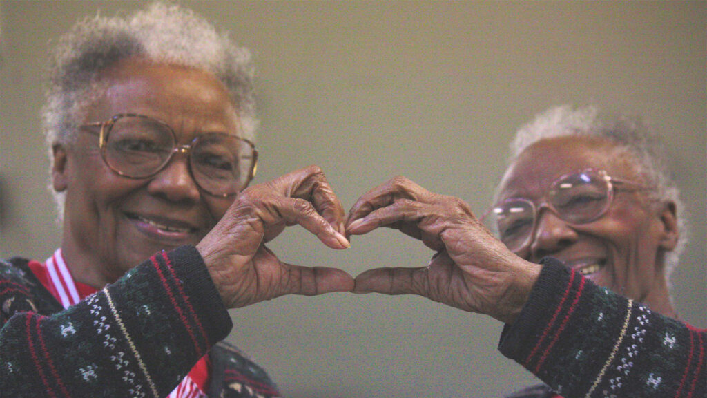 Two elderly women wearing glasses and sweaters smile warmly, forming a heart shape with their hands together. The background is plain and softly lit.