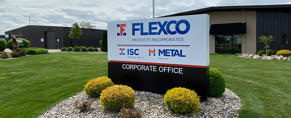 A large sign on a well-manicured lawn reads "FLEXCO PRODUCTS INCORPORATED CORPORATE OFFICE" with ISC and METAL logos. Office buildings and shrubs are visible in the background.