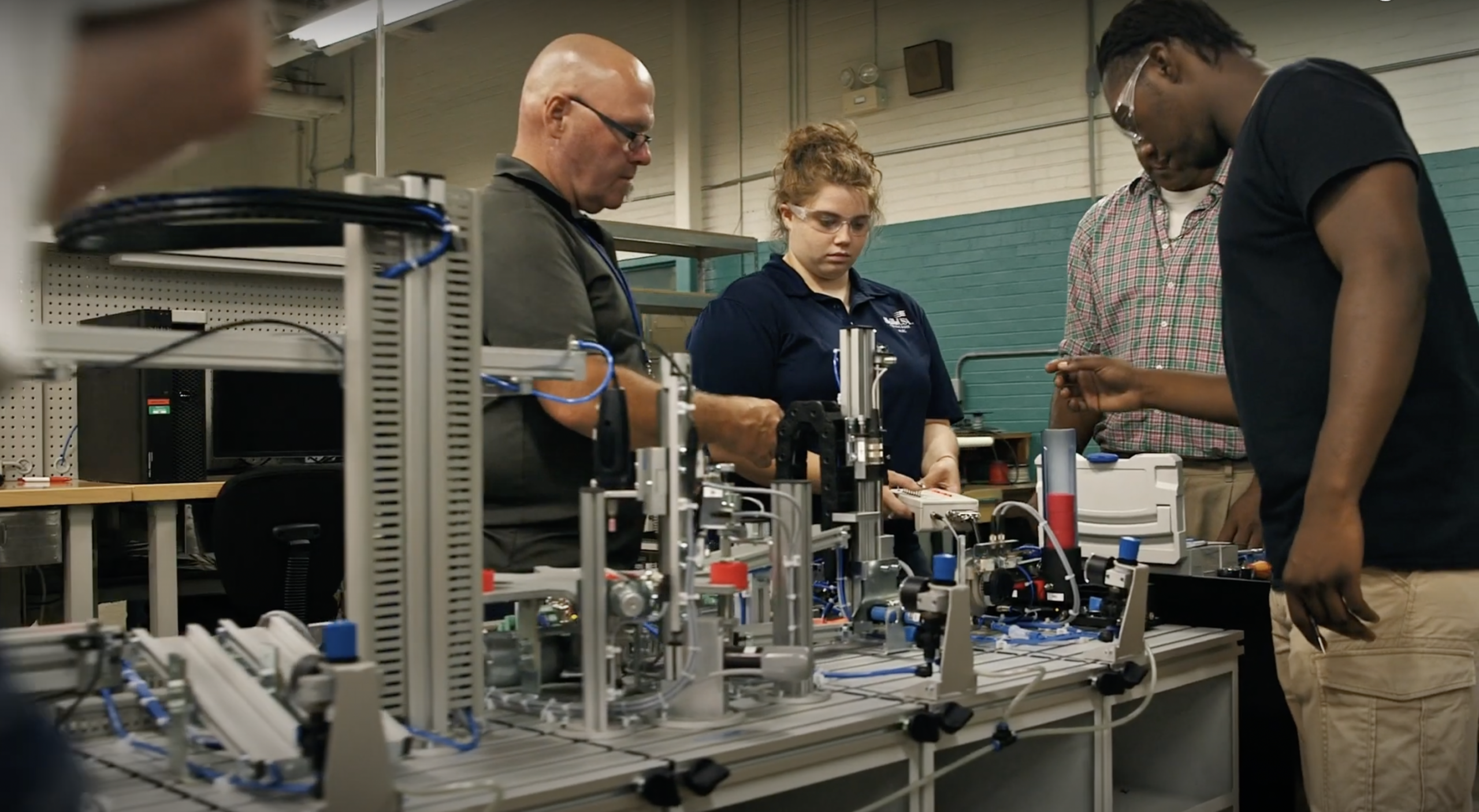 A group of people stand around a table with industrial automation equipment, working together in a lab setting. One man explains something while others observe and participate, all focused on the machinery.