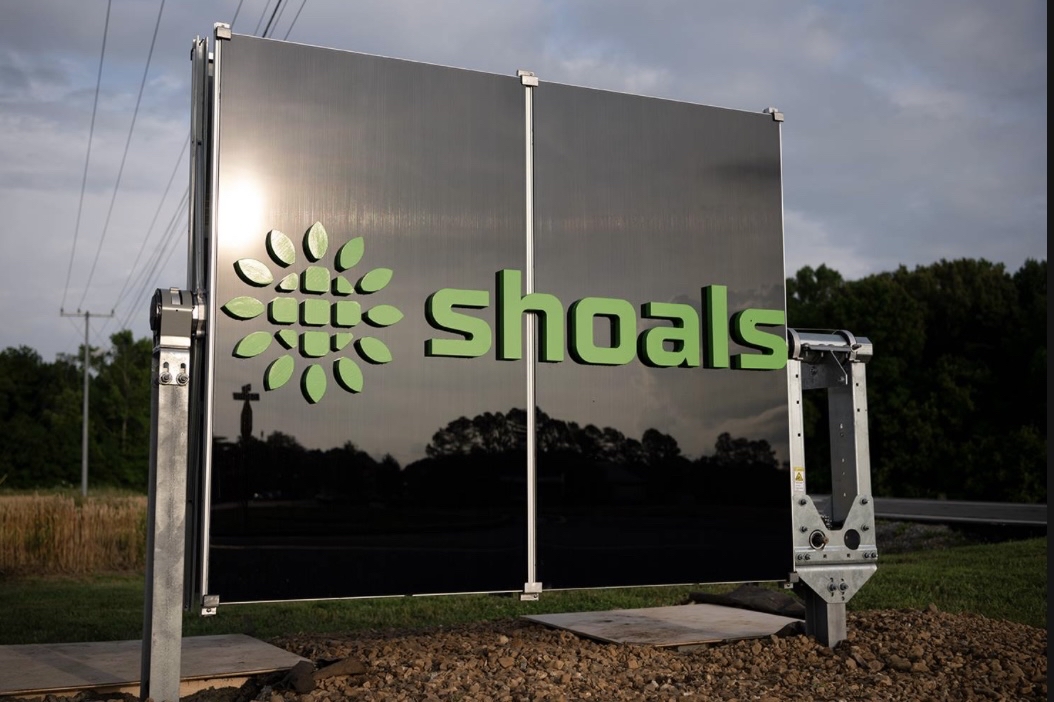 A large black sign with a green logo and the word "shoals" in lowercase letters stands outdoors on gravel, with grass, trees, and a road visible in the background.