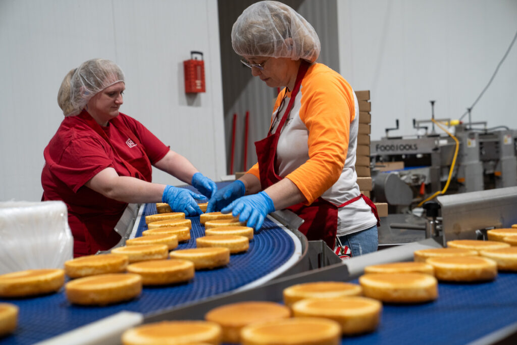 Two workers wearing hairnets, gloves, and aprons arrange round cakes on a conveyor belt in a food processing facility. Machinery and safety equipment are visible in the background.