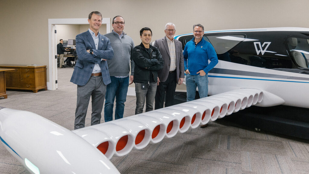 Five men stand smiling beside a futuristic aircraft model with numerous round openings along its wing in a modern office space.