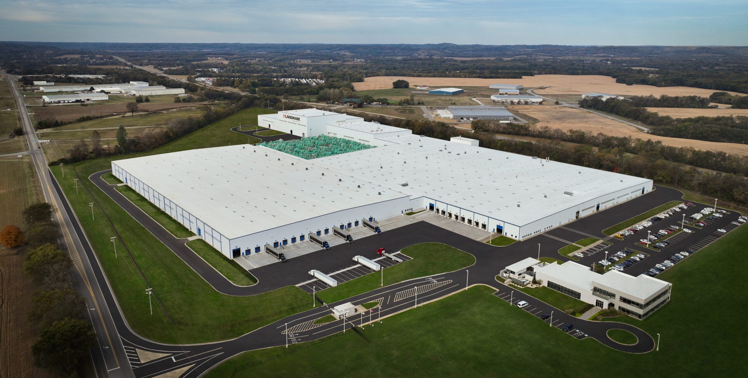 A large white industrial warehouse complex with multiple loading docks and parking lots, surrounded by green lawns and roads, set in a rural landscape with fields and trees under a partly cloudy sky.