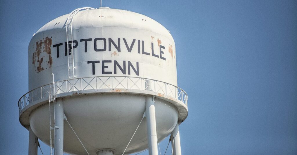 A white water tower with some rust spots displays the words "TIPTONVILLE TENN" against a clear blue sky.