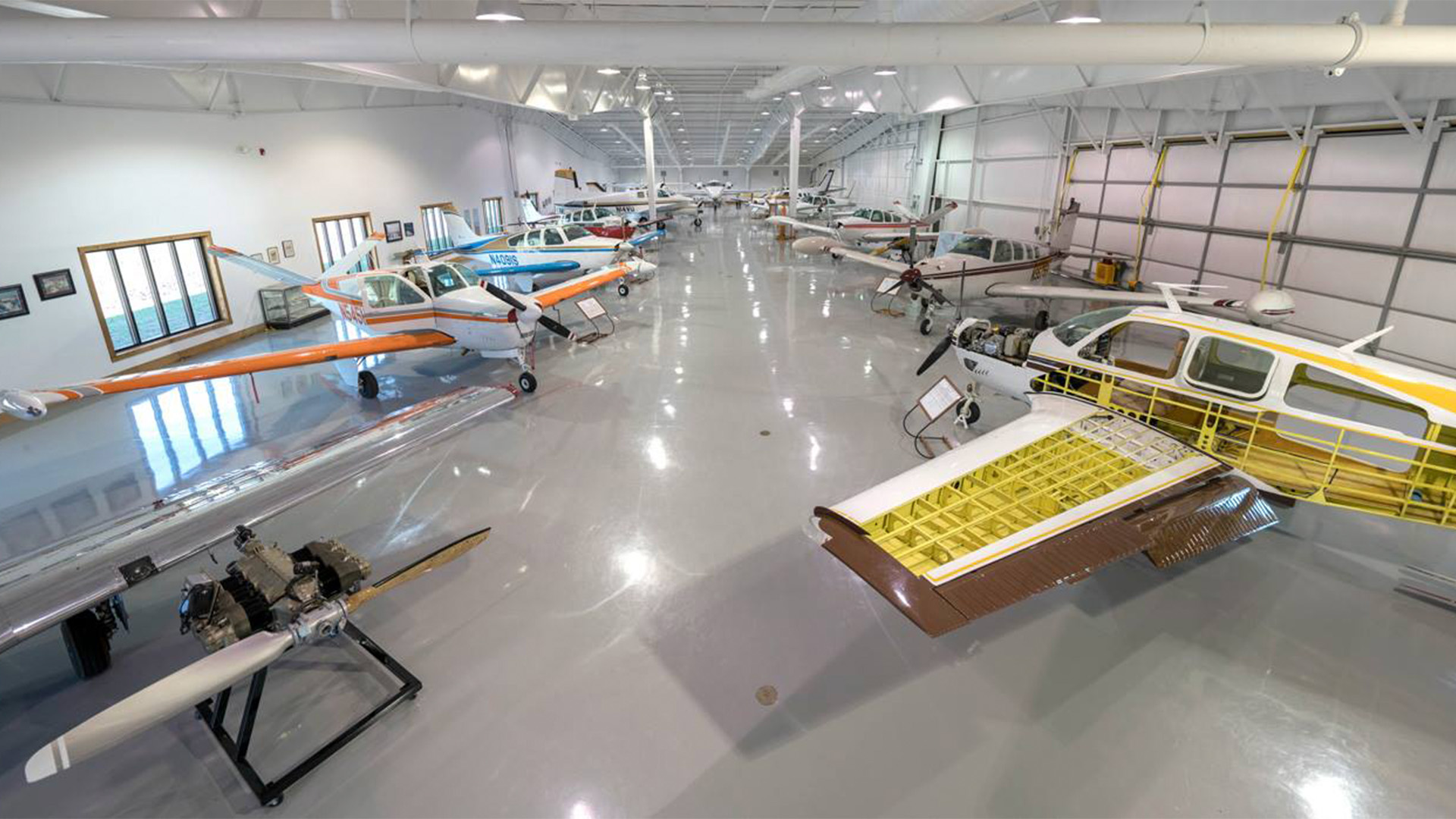 A spacious, well-lit aircraft hangar with several small airplanes parked inside on a shiny, polished floor. Some planes have parts exposed, and the area features high ceilings and large windows.
