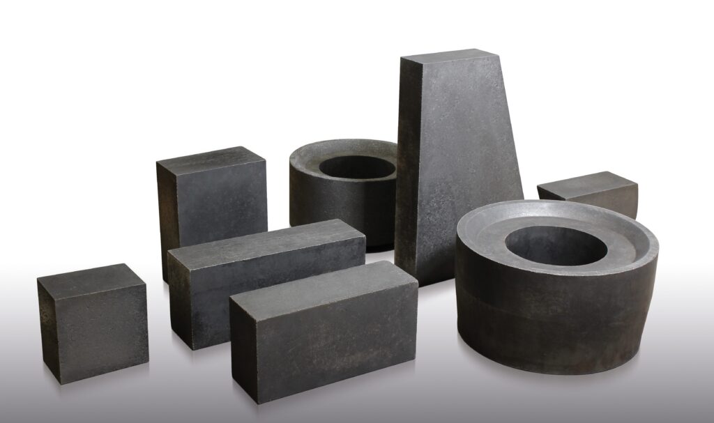 Several dark gray, solid graphite blocks and rings of various shapes and sizes are arranged on a white surface, including rectangular, cylindrical, and trapezoidal forms.