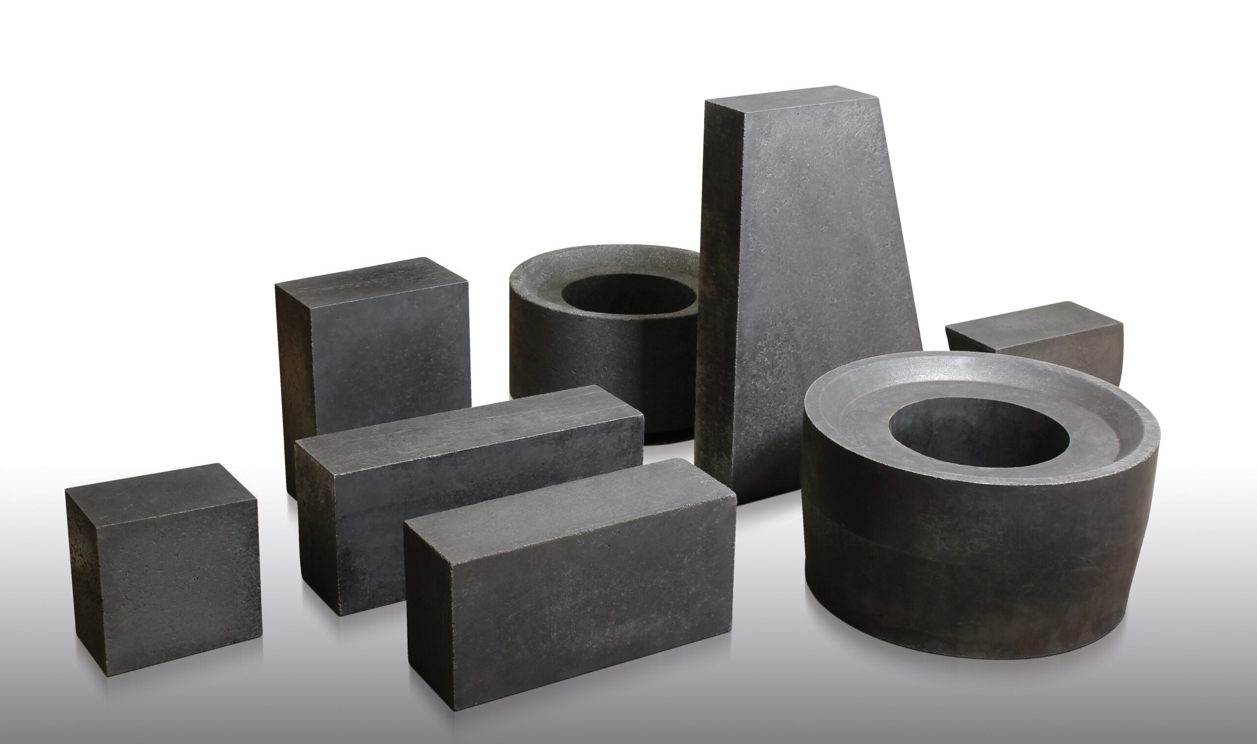 Several dark gray, solid graphite blocks and rings of various shapes and sizes are arranged on a white surface, including rectangular, cylindrical, and trapezoidal forms.