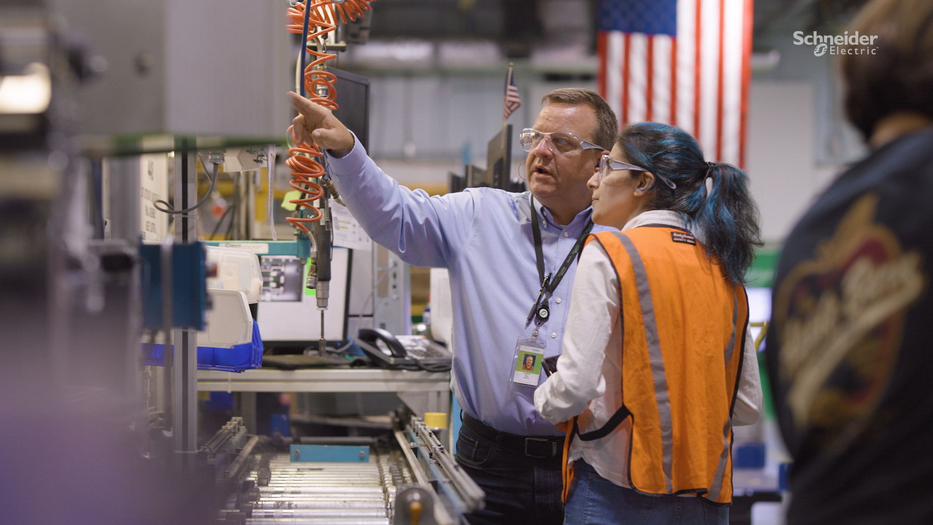 A man in safety glasses points at machinery while explaining something to a woman in a safety vest inside a factory. Equipment, tools, and an American flag are visible in the background.