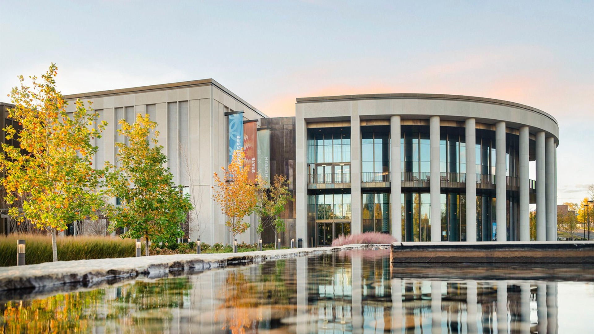 A modern building with large glass windows and tall columns stands beside a reflecting pool, surrounded by trees and landscaping under a partly cloudy sky at sunset.