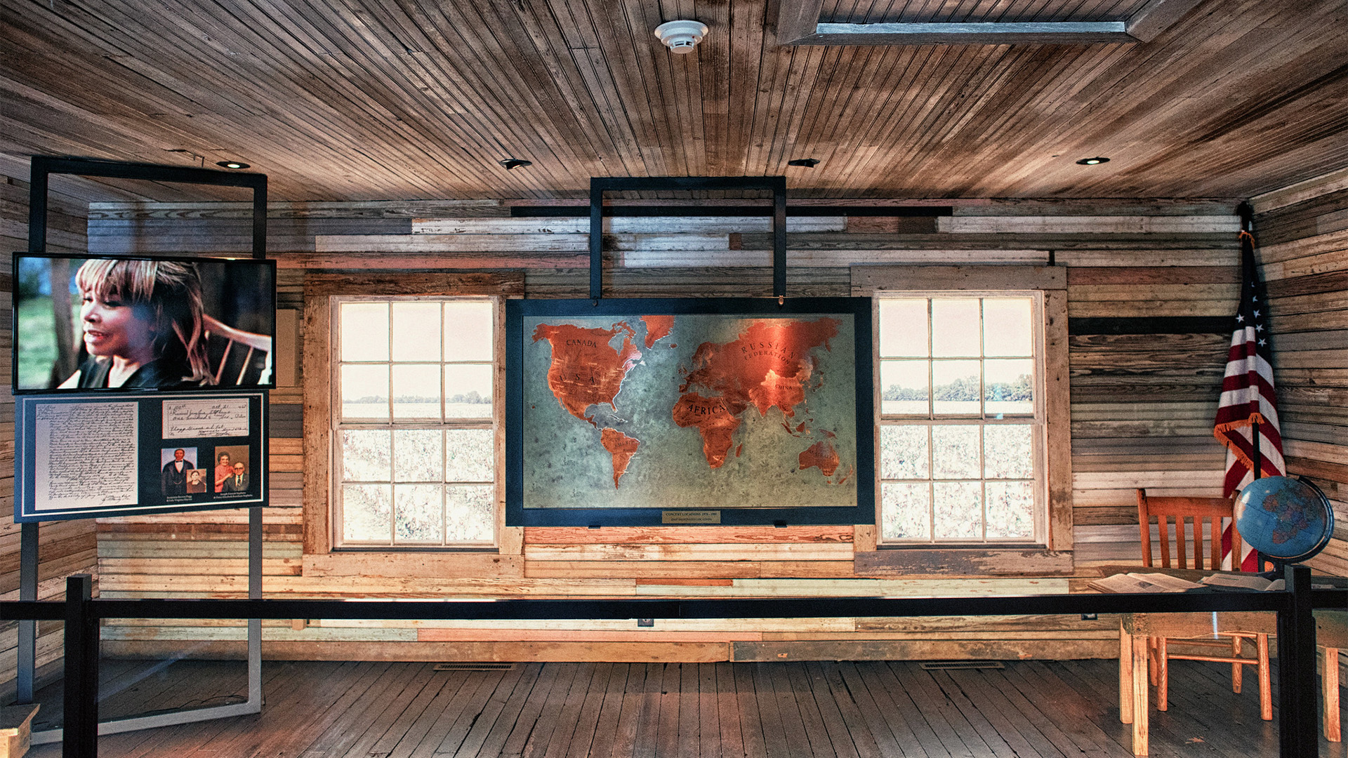 A rustic classroom features a world map between two windows, a TV screen with a woman’s photo and text, an American flag, a globe on a desk with a chair, and wooden walls and floor.