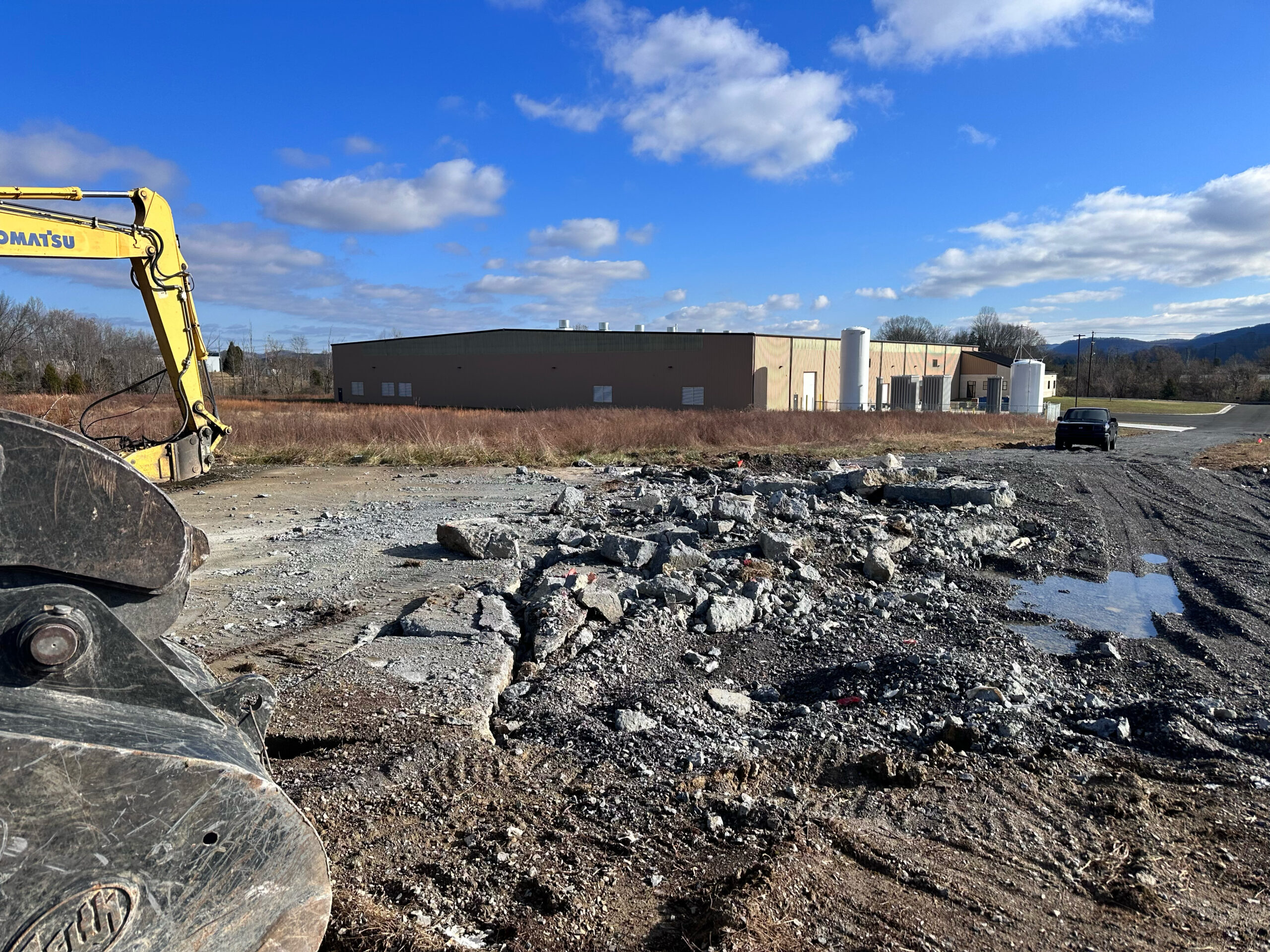 A construction site with broken concrete rubble in the foreground, a yellow excavator on the left, and a beige industrial building in the background under a partly cloudy blue sky.