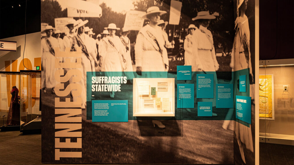 Exhibit display showing a large sepia photo of women suffragists in hats and dresses, with text panels titled “Tennessee” and “Suffragists Statewide,” and historical artifacts displayed in a glass case.