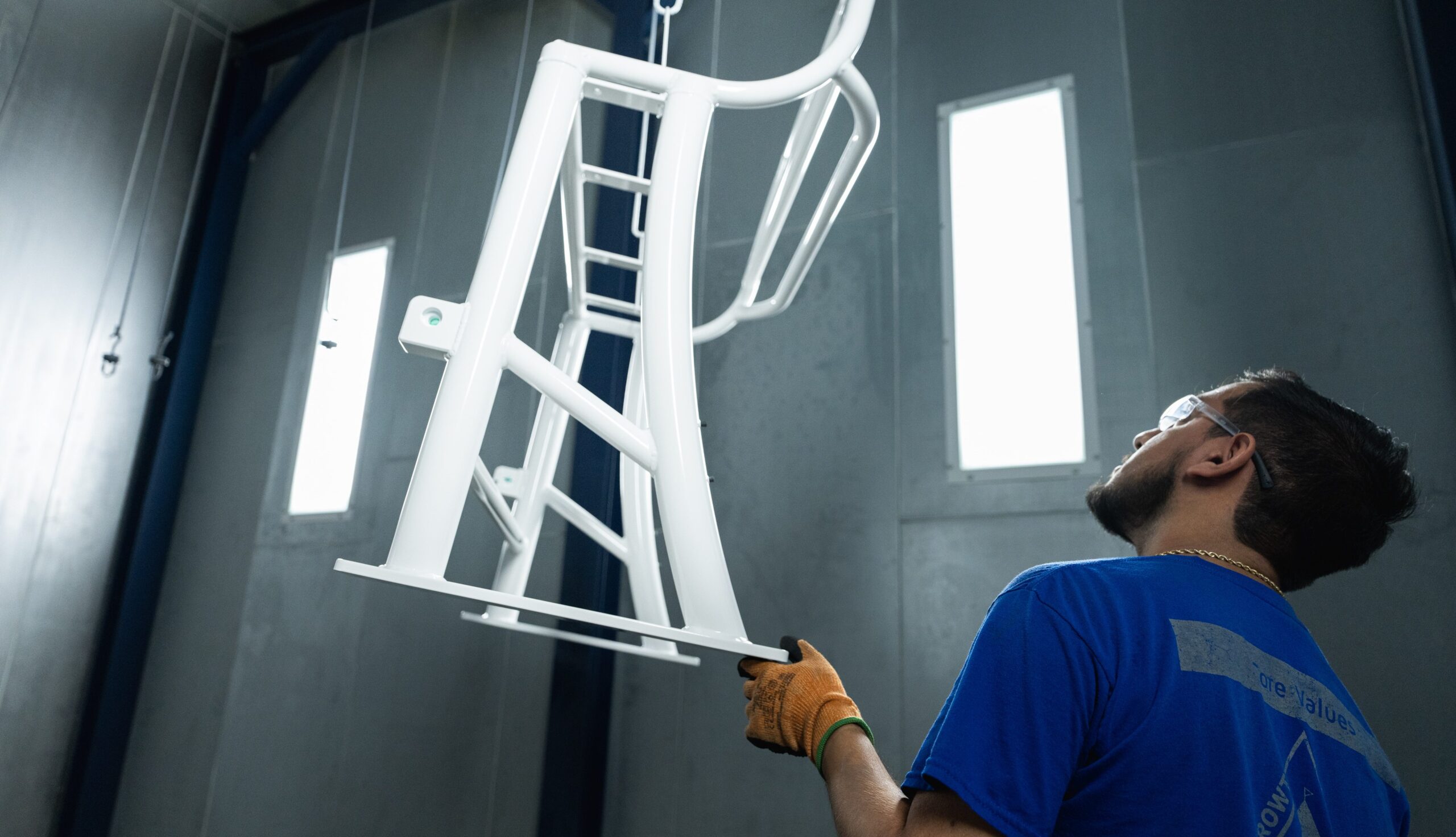 A worker wearing safety glasses and gloves inspects or sprays a large, white metal frame suspended in an industrial room with tall windows and blue walls.