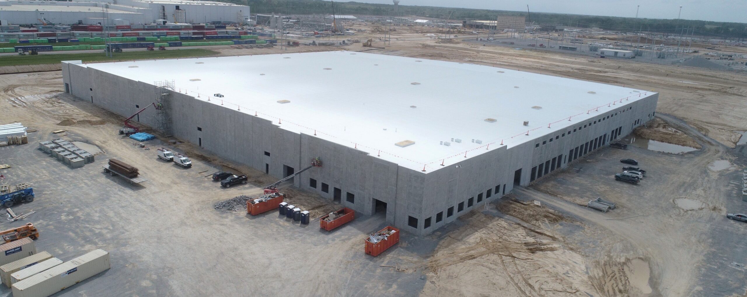 A large, rectangular industrial building under construction sits on a dirt lot, surrounded by construction vehicles, equipment, and materials. The flat white roof contrasts with the gray concrete walls. Nearby areas are cleared and under development.