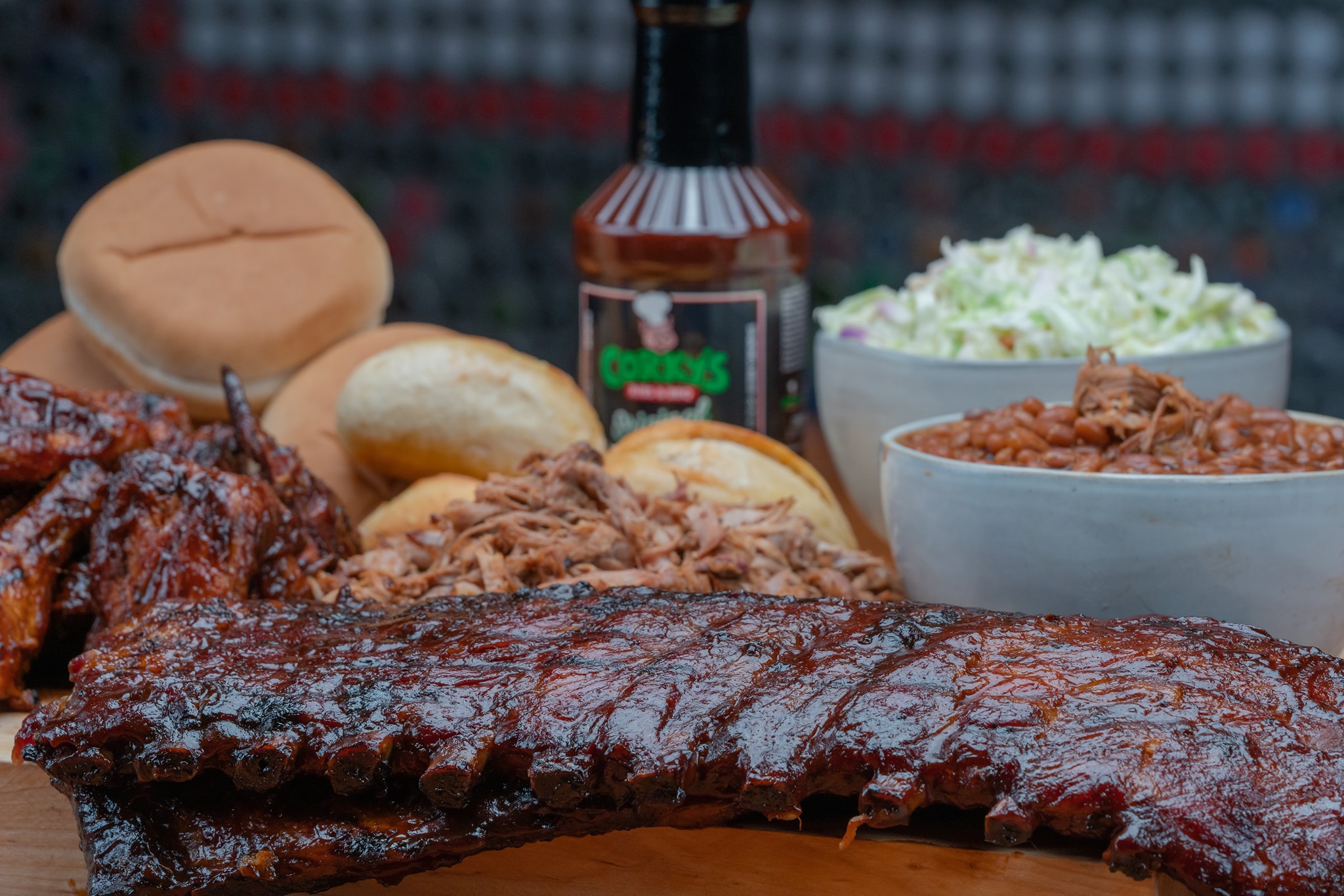 A close-up of a barbecue feast featuring glazed ribs, pulled pork, chicken wings, baked beans, coleslaw, sandwich buns, and a bottle of barbecue sauce, all arranged on a wooden surface.